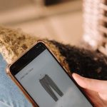 Cross-Border E-commerce - High angle of unrecognizable woman sitting on chair and choosing clothes in internet shop using mobile phone