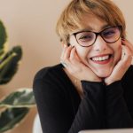 Corporate Retreats - Cheerful woman smiling while sitting at table with laptop