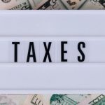 Taxes - Top view of white vintage light box with TAXES inscription placed on stack of USA dollar bills on white surface