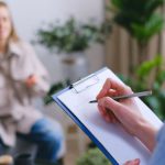 Dilemmas - Unrecognizable professional female psychologist writing on clipboard while sitting against client on blurred background during psychotherapy session in light office